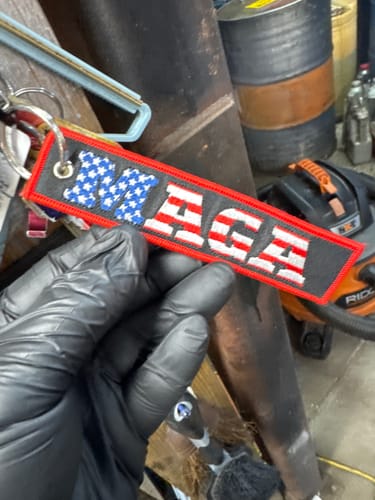A reviewer's gloved hand holding the rectangular MAGA Jet Tag with its American flag lettering.