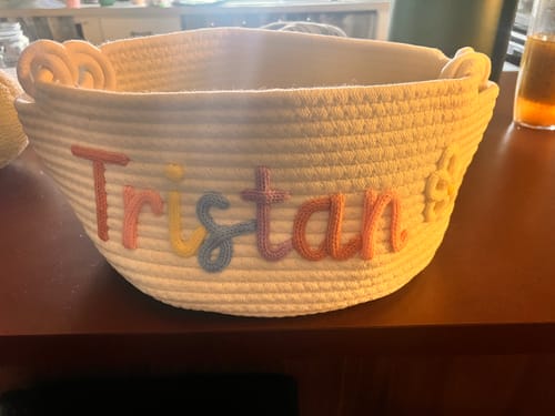 Customer's white personalized knitted basket with the name 'Tristan' in multicolor pastel yarn and a small flower icon.