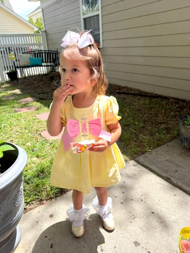 A customer's child wearing the bright yellow Isabella Skirt Bubble with a large pink bow, standing on a patio.