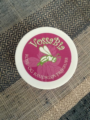Kundens Vossabia Rose- og raudkløverskrubb i en hvit, rund krukke på et mønstret stoff.