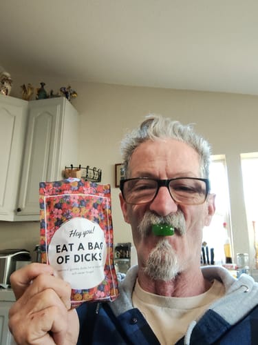Reviewer holding the cheeky Bag Of Gummy Candy D's and posing with a green gummy balanced on their lip.