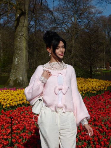 Kundin trägt den rosa Twist & Schleife Wendepullover offen vor einem bunten Tulpenfeld.