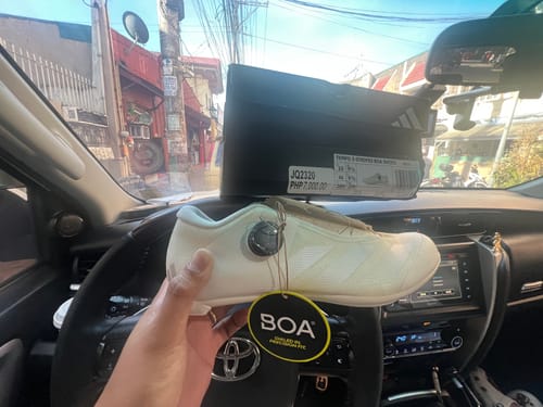 Reviewer holds a white adidas Tempo Boa cycling shoe inside a car, with the shoebox visible in the background.