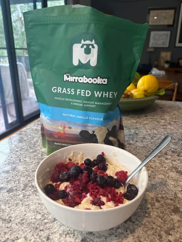 Reviewer's Mirrabooka Grass Fed Whey pouch on a kitchen counter beside a prepared protein bowl with fresh berries.