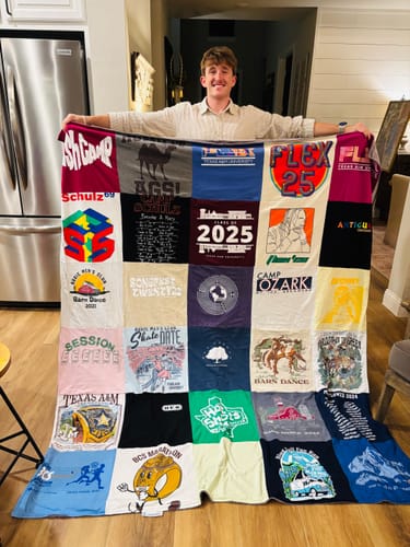 A person proudly holds up a large, multi-colored T-Shirt Quilt made of various memory shirts in a home setting.
