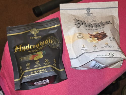 Customer's bags of Hydroglyph fasting formula and Planta plant protein from the Mike Rashid Birthday Bundle on a pink surface.