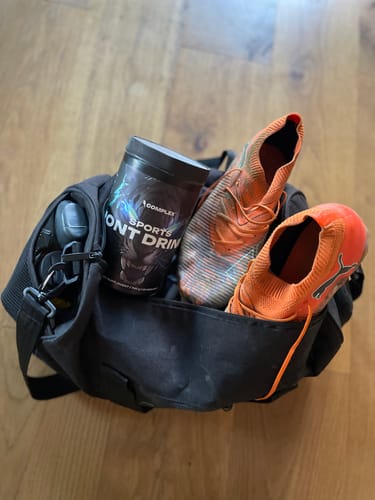 Customer's tub of Sports Iont Drink packed in a black sports bag with a pair of orange soccer cleats.