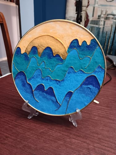 Reviewer's finished craft project from the subscription box, a circular art piece of blue mountains and a gold sun on a stand.