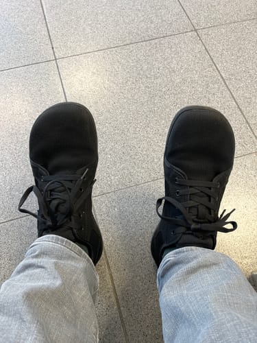 Ein Kunde trägt die schwarzen Luck Ⅰ - Barfußschuhe mit breiter Zehenbox auf einem gefliesten Boden.
