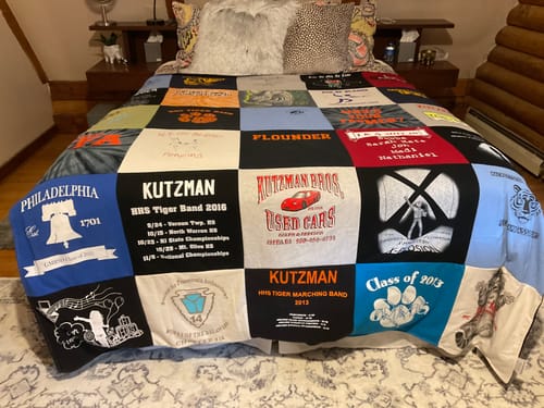 Customer's colorful Premium T Shirt Quilt displayed on a bed, showing patches from a high school band and local businesses.