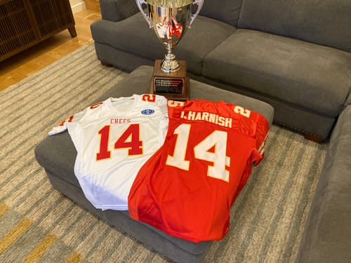 Customer's two custom football jerseys, one white and one red, displayed on an ottoman in front of a trophy.