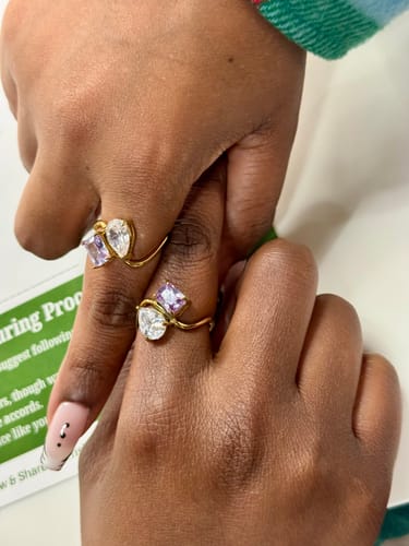 Two people showing their matching Navae Birthstone Rings, with clear pear-cut and purple radiant-cut stones on gold bands.