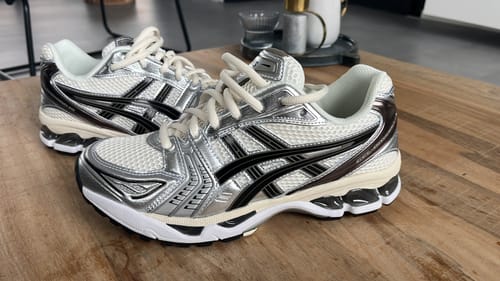 Een recensent toont de ASICS Gel-Kayano 14 sneakers in crème, zilver en zwart op een houten tafel.