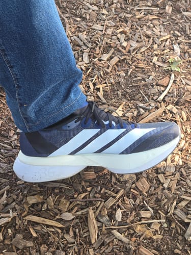 Customer wearing a dark blue Adizero Boston 13 Running Shoe with light blue stripes and a thick white sole outdoors.