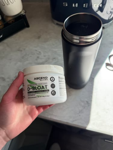 Customer holding a white tub of D-BLOAT on a kitchen counter next to a black travel mug filled with the prepared drink.