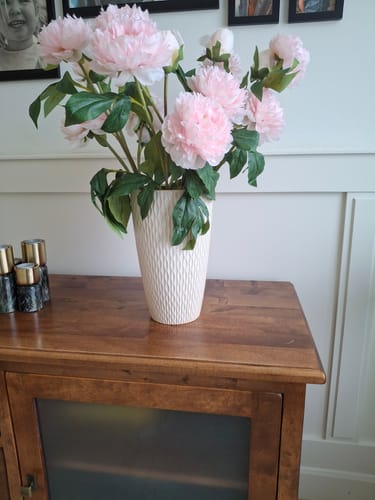 Recensent toont de volle, roze Zijden Pioenrozen in een witte vaas op een houten dressoir.