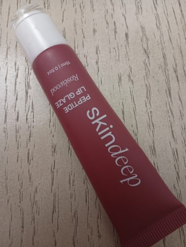 Customer's Peptide Lip Glaze tube in the shade Roséwood resting on a light wood surface.