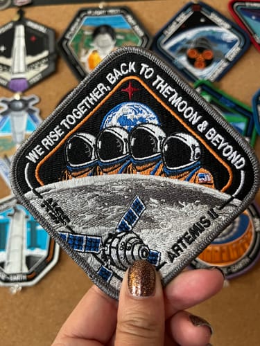 A reviewer's hand holding the detailed Artemis II - Collectors Patch in front of a board of other space patches.