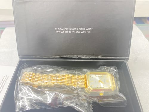 Close-up of the gold CLAIRE DE MAINS watch in protective plastic inside its black presentation box.
