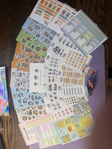 Customer's view of the numerous sticker sheets from their Oopsie Mystery Bag laid out on a wooden surface.