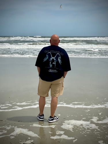 Customer on a beach showing the back design of the black Galatians 5:1 oversized t-shirt.