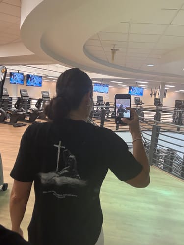 Reviewer at the gym taking a mirror selfie to show the back design of the 'Warrior's Prayer' T-Shirt.