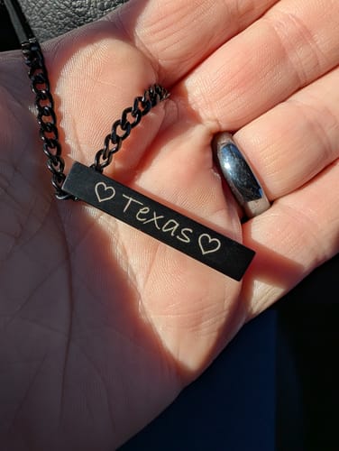Customer's hand holding a matte black bar urn pendant engraved with the name 'Texas' between two hearts.