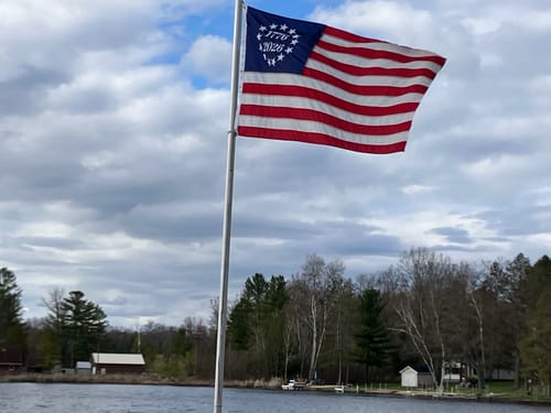 Reviewer's 1776 to 2026 Betsy Ross Flag flying on a flagpole over a lake under a cloudy sky.
