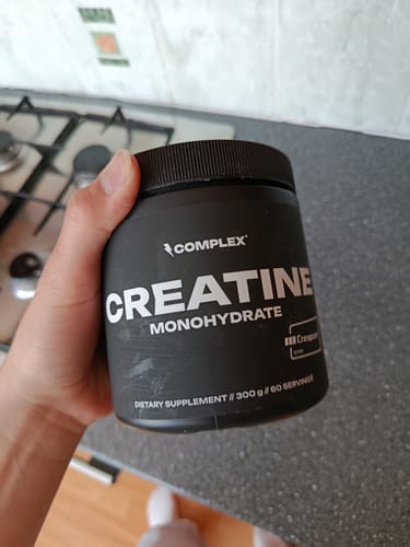 Reviewer holding the black tub of Complex Creatine Monohydrate supplement in a kitchen.