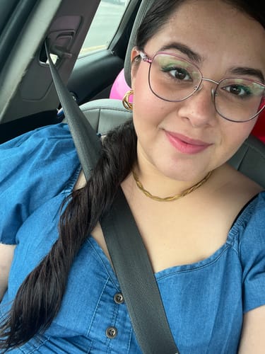 Una clienta sonríe mientras lleva el Collar Olga Triple dorado con una blusa azul en un coche.