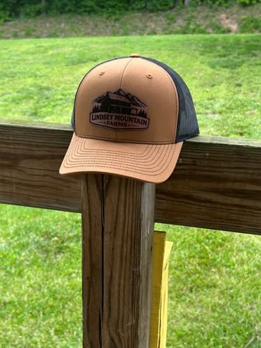 Customer's brown and black custom leather patch trucker hat resting on a wooden fence post outdoors.