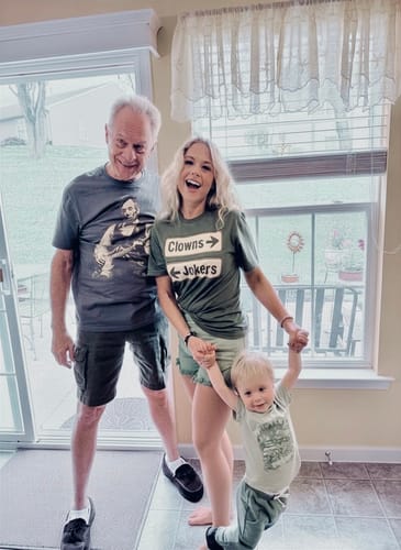 Reviewer wearing a green Clowns or Jokers T-Shirt, laughing while standing indoors with her father and a child.