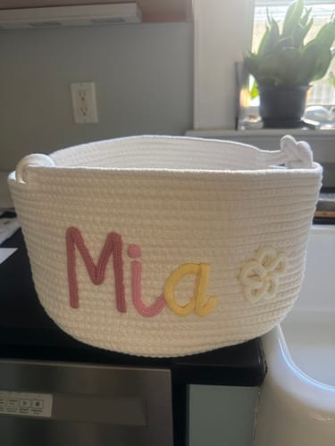 Customer's personalized white woven basket with the name 'Mia' in pink and yellow letters and a white flower icon.