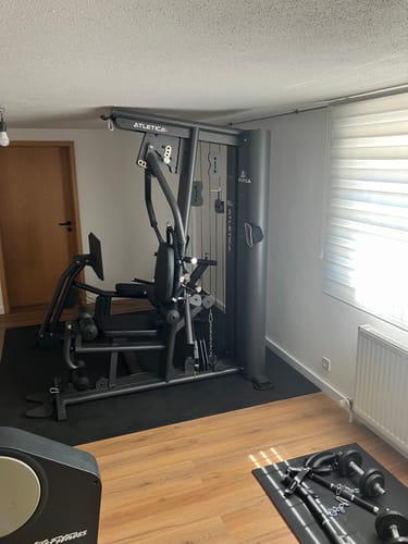 Ein Kunde zeigt das aufgebaute X-Tron Multigym mit Beinpresse, das auf einer schwarzen Matte in einem Raum mit Holzboden steht.