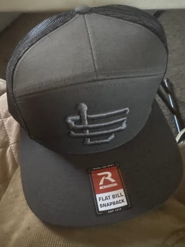 Customer's dark gray Stealth Seven Panel Trucker Cap, showing the front LS logo and flat bill with a sticker.