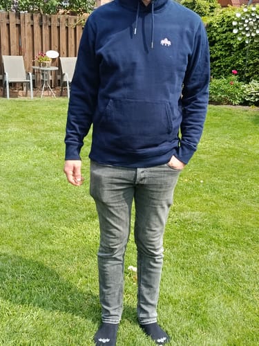 Kunde trägt den blauen Holzbrüder Stick Bio-Hoodie mit lockerer Passform in einem Garten.