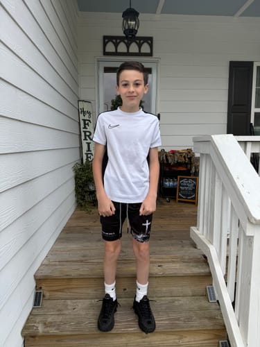 Customer wearing the black 'Cross' Youth Performance Shorts on a porch, showing a full view of the product's fit and design.