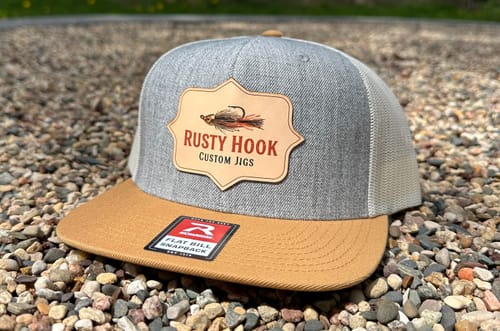 Customer's grey and tan flatbill trucker hat with a custom leather patch, shown outdoors on a gravel surface.