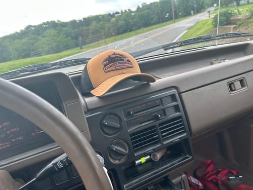 Reviewer's brown and black Custom Leather Patch Trucker Hat resting on a car dashboard.