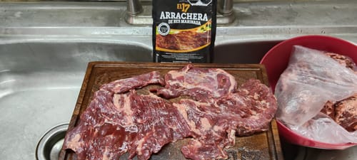 Cliente mostrando la Arrachera Marinada cruda sobre una tabla de cortar, con el empaque del producto al fondo.