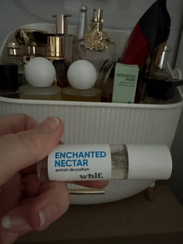 Reviewer holding a small vial of Enchanted Nectar perfume with a collection of other fragrances in the background.