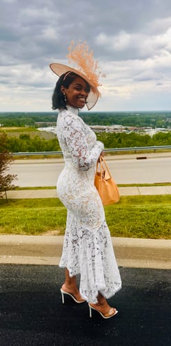 Customer smiling outdoors in her white Ishani Lace Midi Dress, styled with a large fascinator hat.