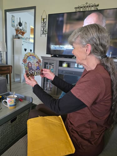 A woman sits in a living room, smiling as she holds the personalized sunflower suncatcher.