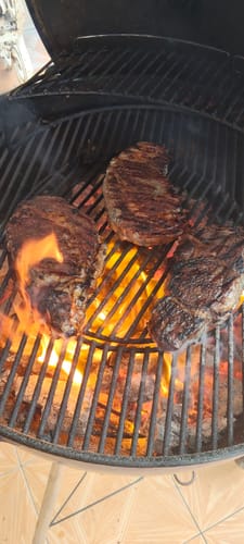 Cliente cocinando tres filetes de Rib Eye Steak Prime en una parrilla de carbón sobre llamas.
