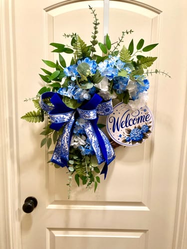 Customer's 14" Grapevine Wreath decorated with blue hydrangeas, green foliage, and a large blue patterned bow.