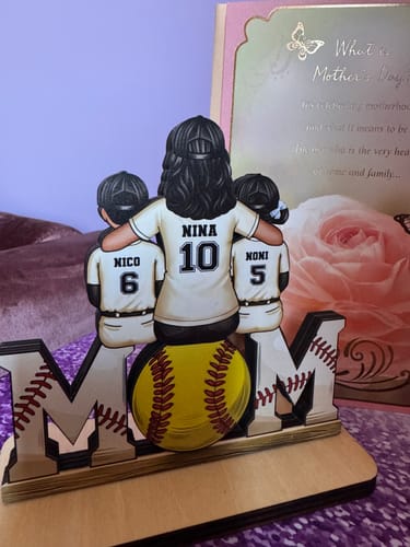 Customer's personalized wooden plaque of a mom and two kids in jerseys, sitting on the word 'MOM' with a softball for the 'O'.