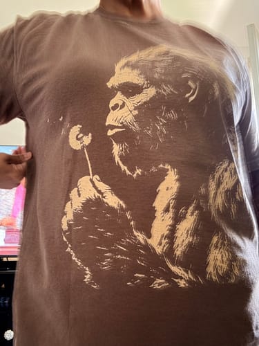 A reviewer wearing the brown Blowing Wishes T-Shirt, which features a large graphic of a sasquatch blowing on a dandelion.