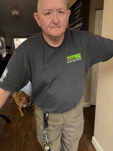 Customer wearing the gray T.V.P. Elite Membership t-shirt with a green logo on the front in a home setting.
