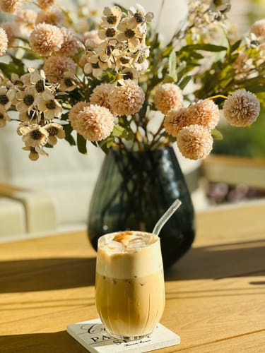 Close-up van het Wax Arrangement - Crème van een klant in een donkere vaas naast een ijskoffie op tafel.