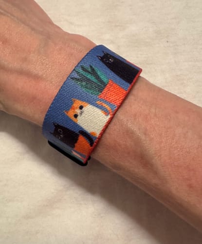 Customer's wrist wearing the colorful Handle With Care Watchband+ featuring a ginger cat and black cats.